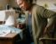 An Black American woman holding her lower back in pain while sitting at a sewing machine in her home studio.