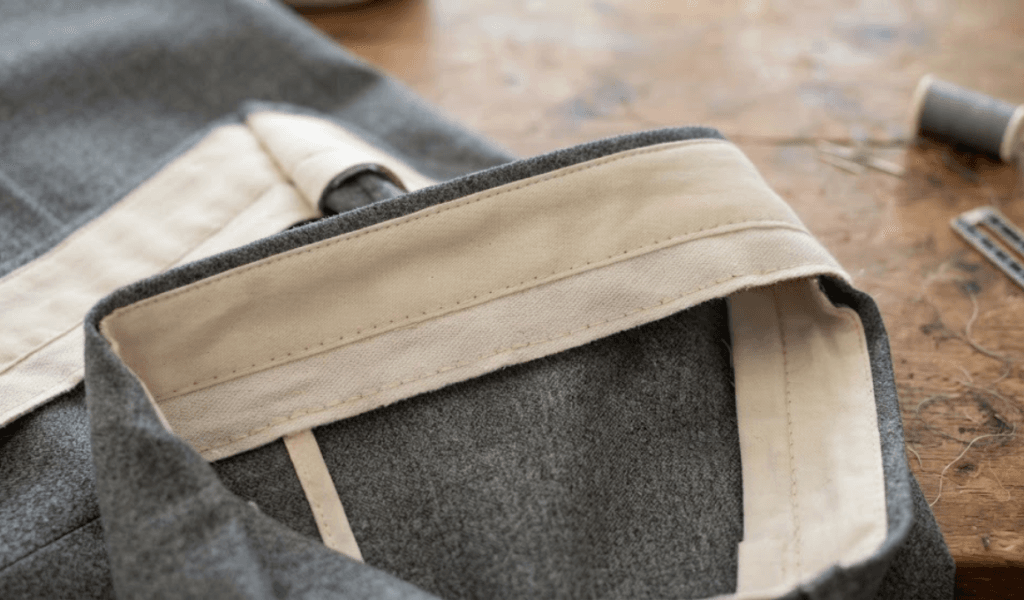 Close-up of a tailored trouser waistband under construction, showing the cream interfacing and facing folded back to reveal the interior stitching against gray wool fabric.