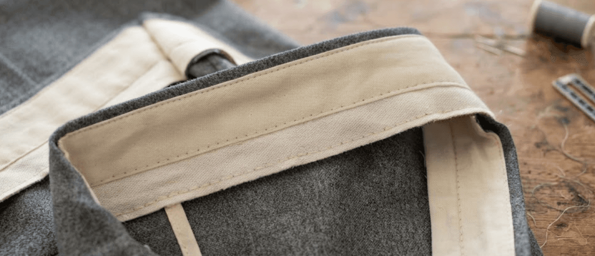 Close-up of a tailored trouser waistband under construction, showing the cream interfacing and facing folded back to reveal the interior stitching against gray wool fabric.
