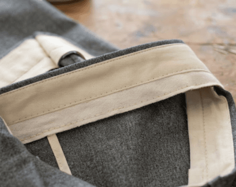 Close-up of a tailored trouser waistband under construction, showing the cream interfacing and facing folded back to reveal the interior stitching against gray wool fabric.